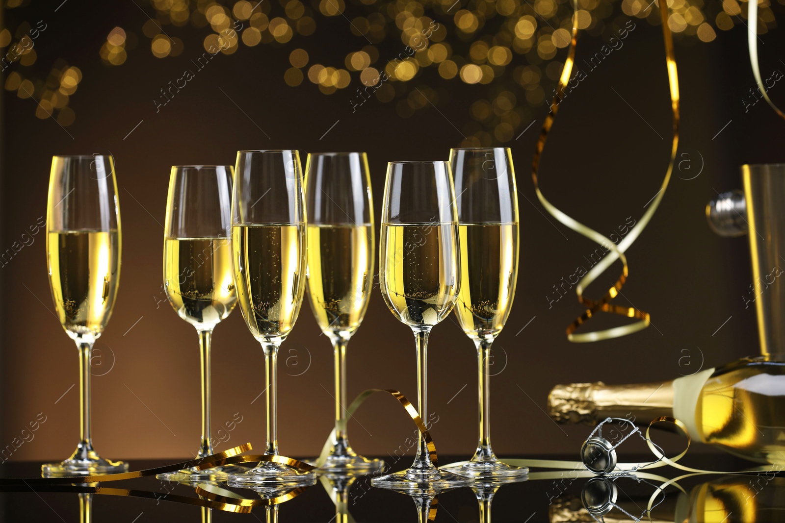 Photo of Champagne in glasses and bottle on mirror surface against brown background with blurred lights