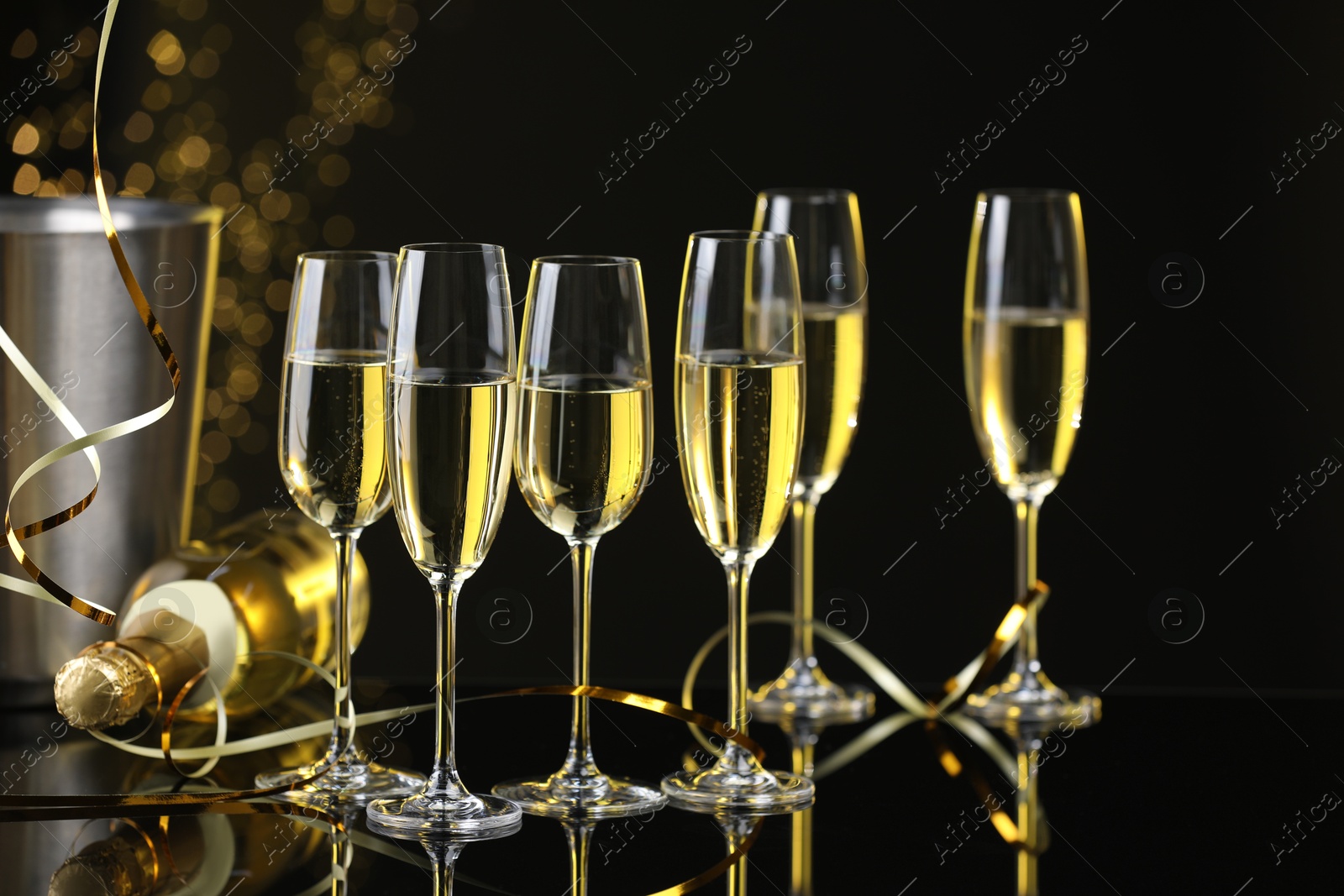 Photo of Champagne in glasses and bottle on mirror surface against black background with blurred lights