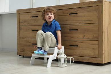 Photo of Little boy sitting on step stool near chest of drawers at home