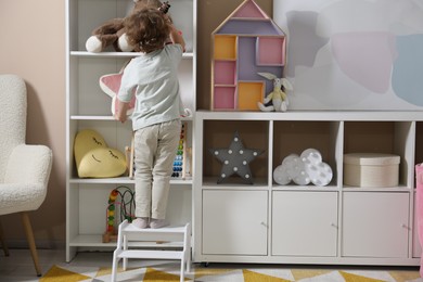 Photo of Little boy standing on step stool and reaching for toys at home, back view