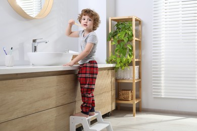 Photo of Little boy brushing teeth while standing on step stool near bathroom vanity indoors