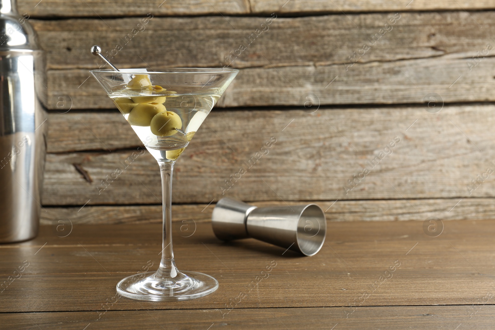 Photo of Martini cocktail with olives in glass and jigger on wooden table, space for text