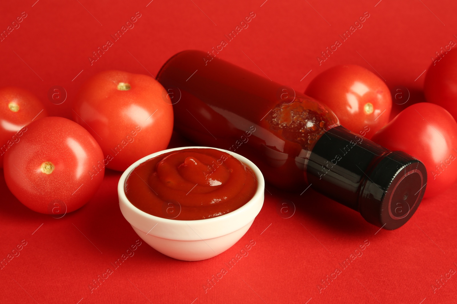 Photo of Ketchup in glass bottle, bowl and fresh tomatoes on red background