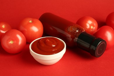 Photo of Ketchup in glass bottle, bowl and fresh tomatoes on red background