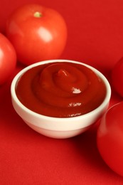 Photo of Ketchup in bowl and fresh tomatoes on red background, closeup