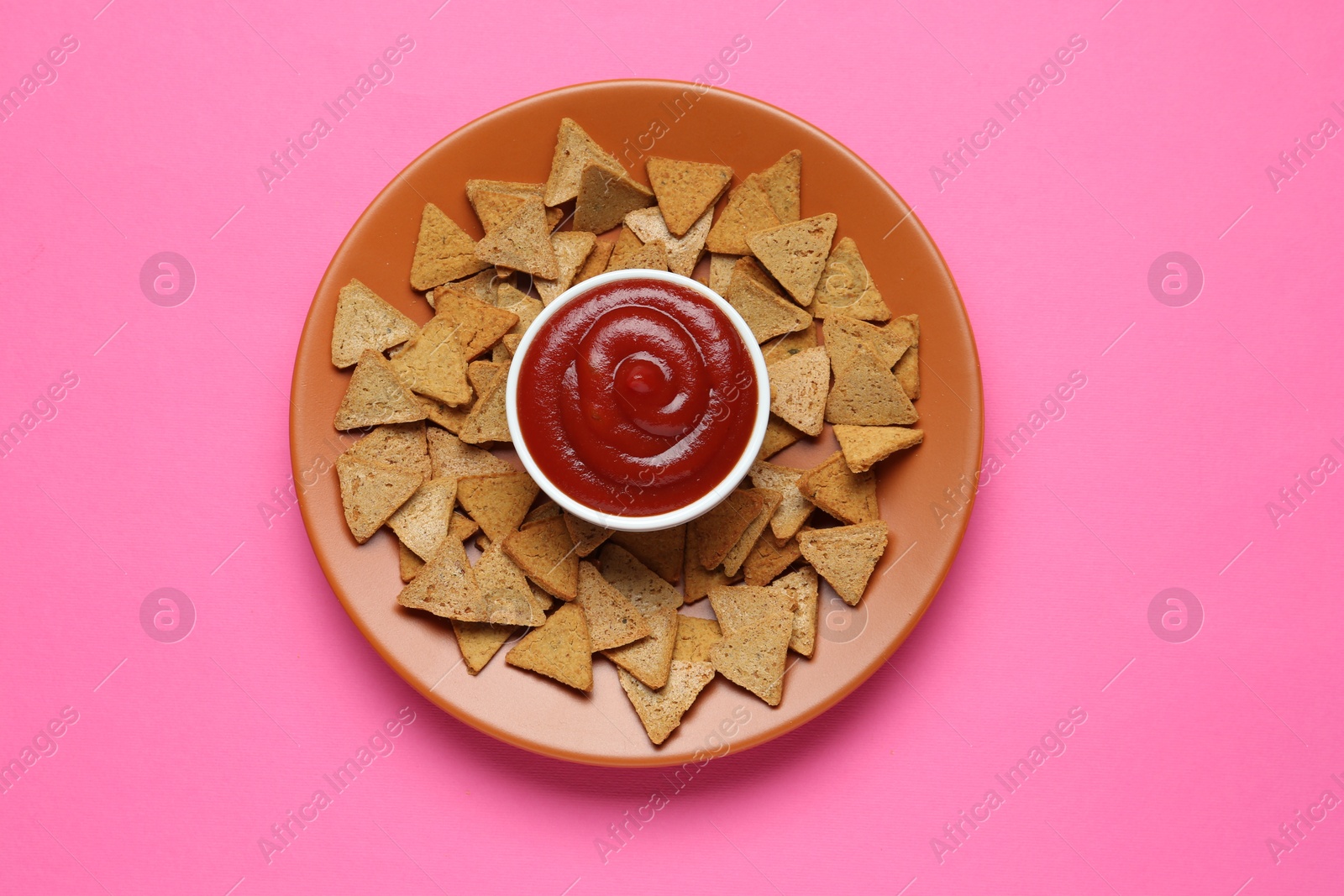 Photo of Ketchup in bowl and pieces of dry bread on pink background, top view