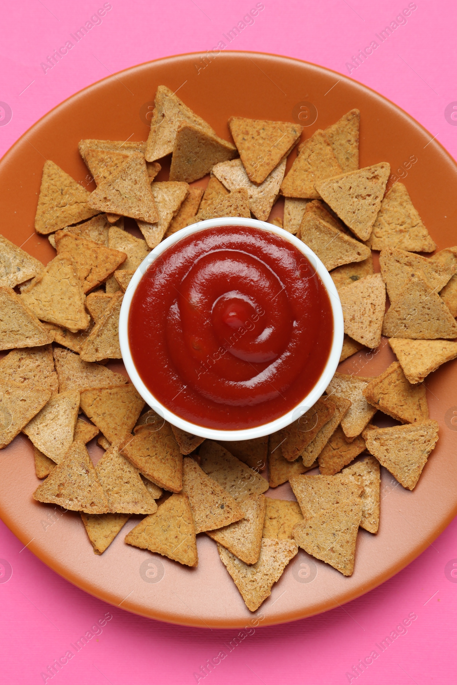 Photo of Ketchup in bowl and pieces of dry bread on pink background, top view