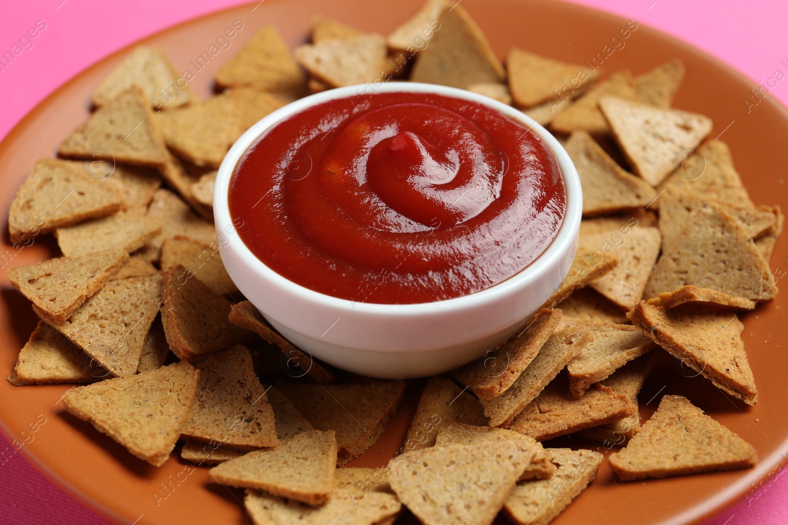 Photo of Ketchup in bowl and pieces of dry bread on pink background, closeup