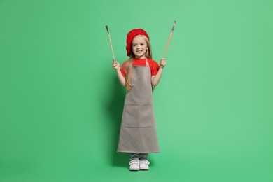 Photo of Little girl with brushes pretending to be artist on green background. Dreaming of future profession