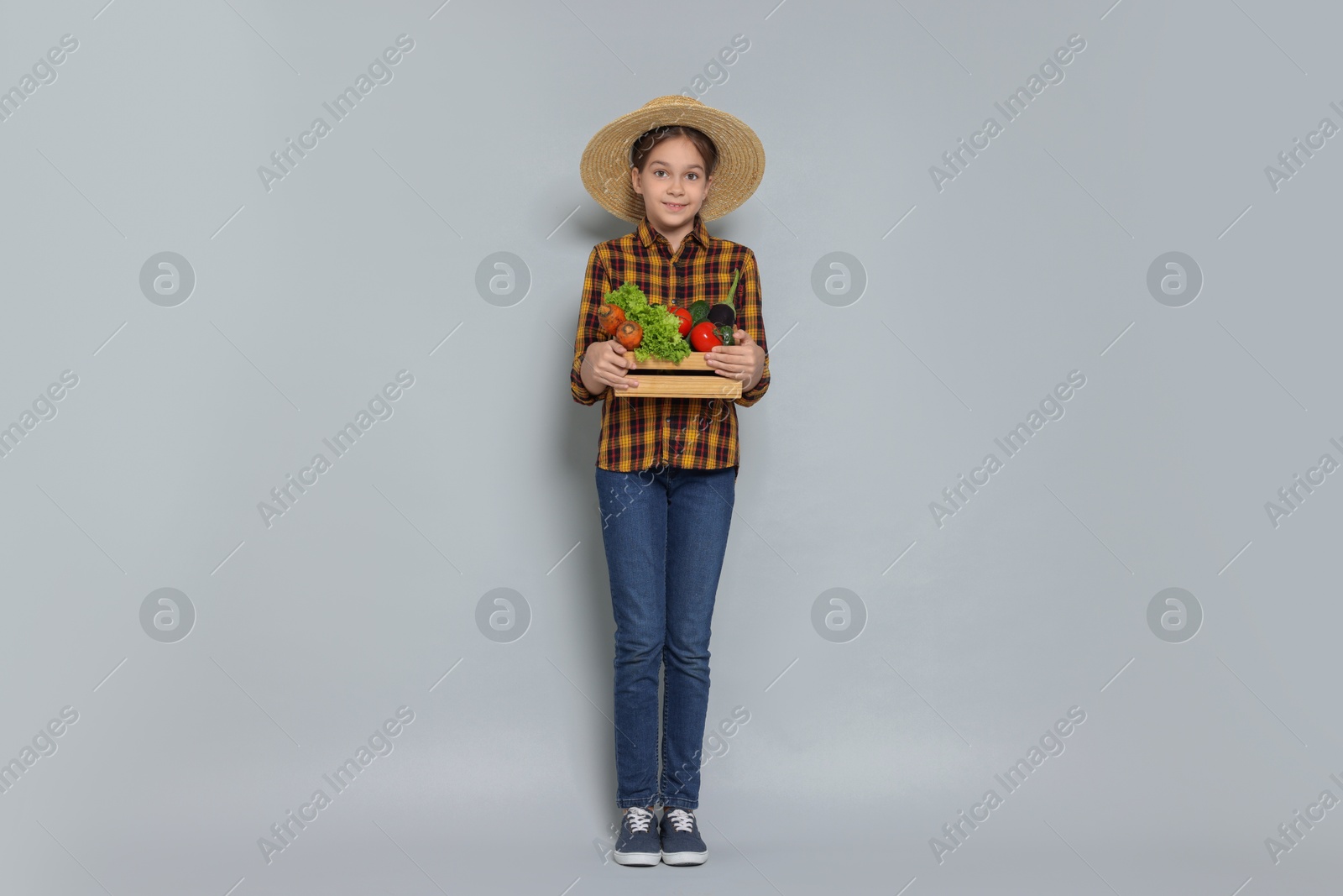 Photo of Girl with straw hat, wooden crate and vegetables pretending to be farmer on light grey background. Dreaming of future profession