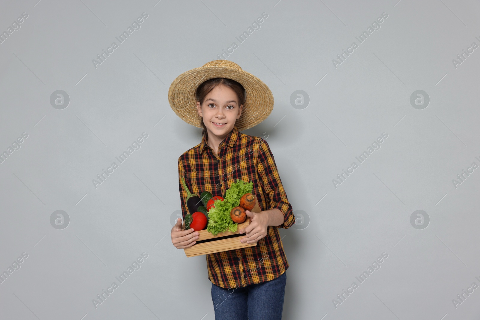 Photo of Girl with straw hat, wooden crate and vegetables pretending to be farmer on light grey background. Dreaming of future profession