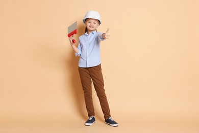 Photo of Little girl in hard hat with putty knife on beige background. Dreaming about future profession