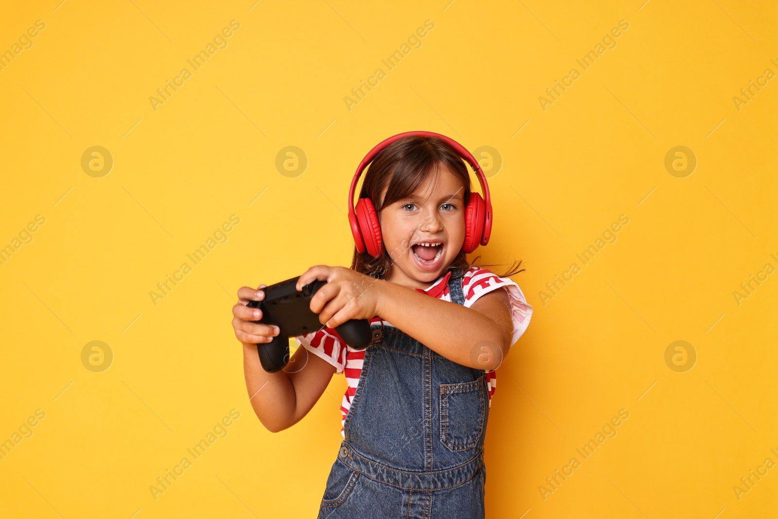 Photo of Happy little girl in headphones playing video game with controller on orange background