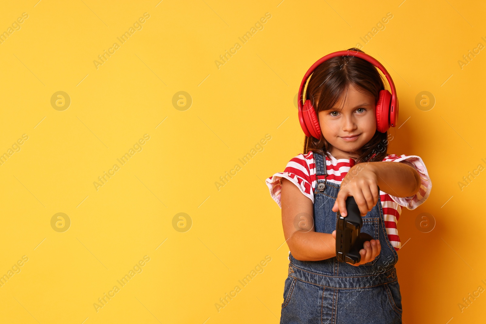 Photo of Cute little girl in headphones playing video game with controller on orange background. Space for text