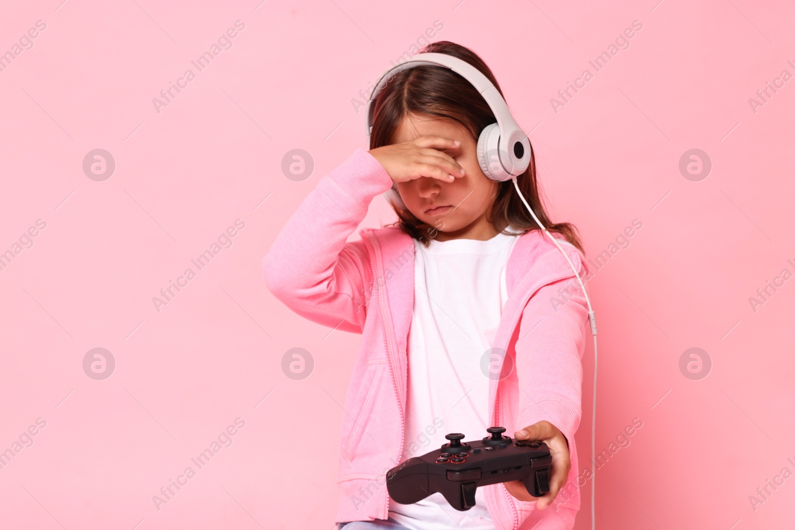 Photo of Little girl in headphones with controller covering eyes on pink background. Space for text