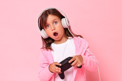 Photo of Cute little girl in headphones playing video game with controller on pink background