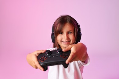 Photo of Happy little girl in headphones playing video game with controller on pink background