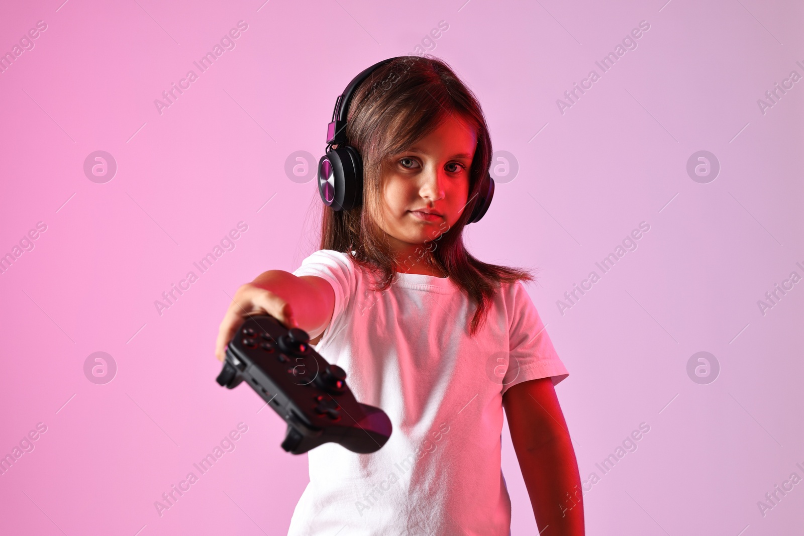 Photo of Cute little girl in headphones showing controller on pink background