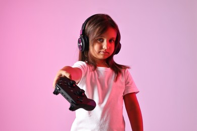 Photo of Cute little girl in headphones showing controller on pink background