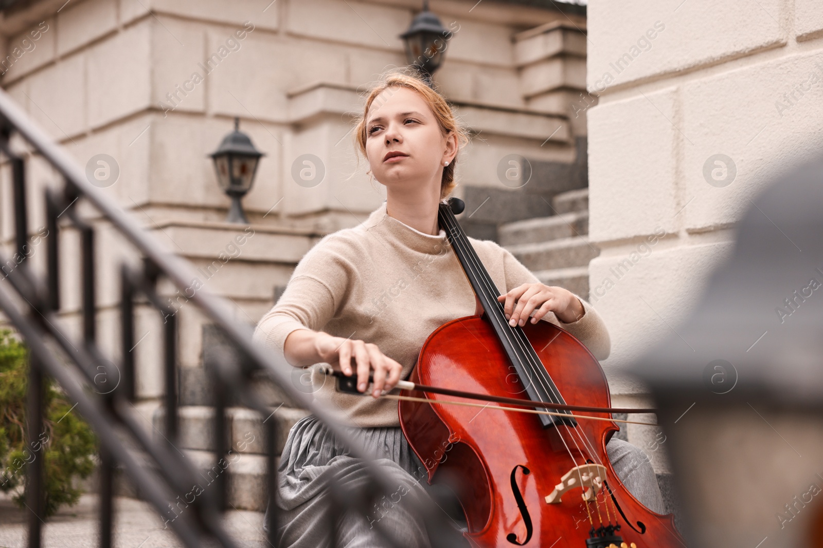 Photo of Beautiful young woman playing cello on stairs outdoors. Classic musical instrument