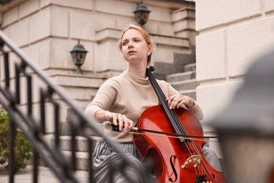 Photo of Beautiful young woman playing cello on stairs outdoors. Classic musical instrument