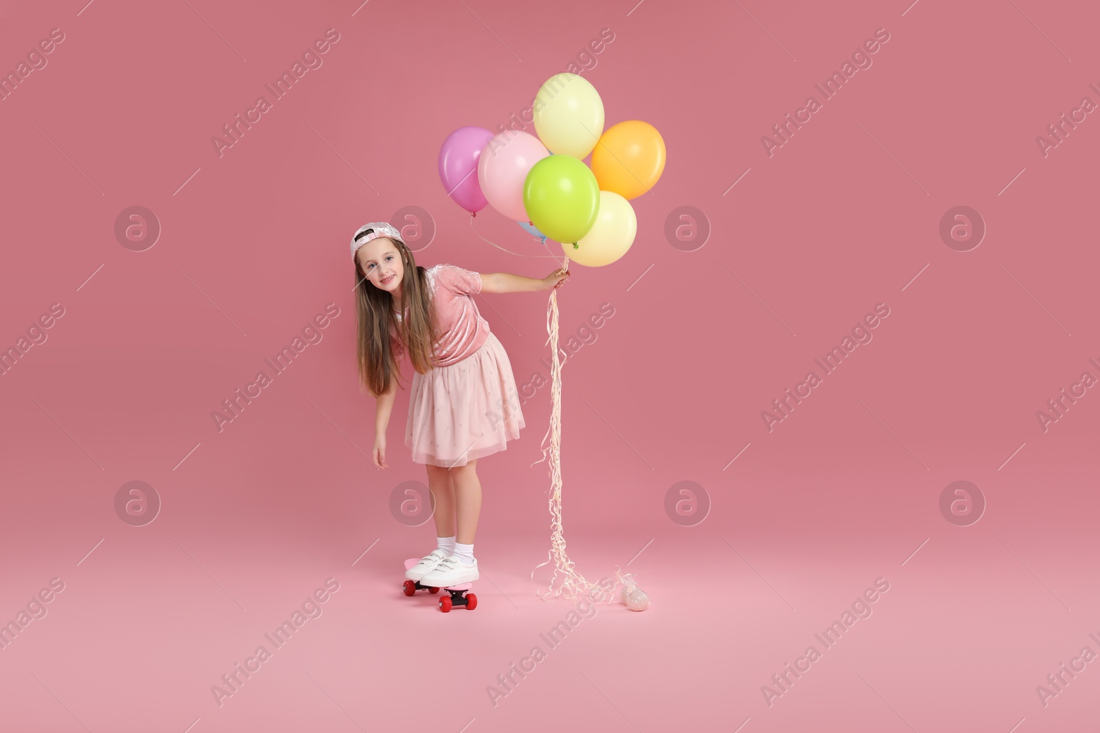 Photo of Little girl with colorful balloons standing on penny board against pink background