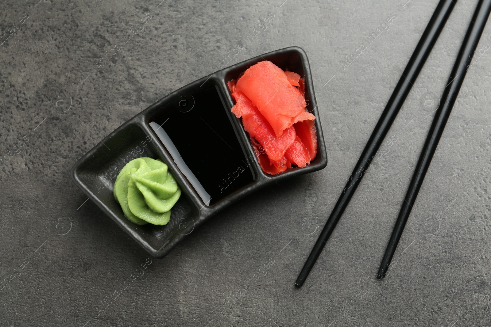 Photo of Hot wasabi paste, soy sauce, ginger and chopsticks on grey textured table, top view