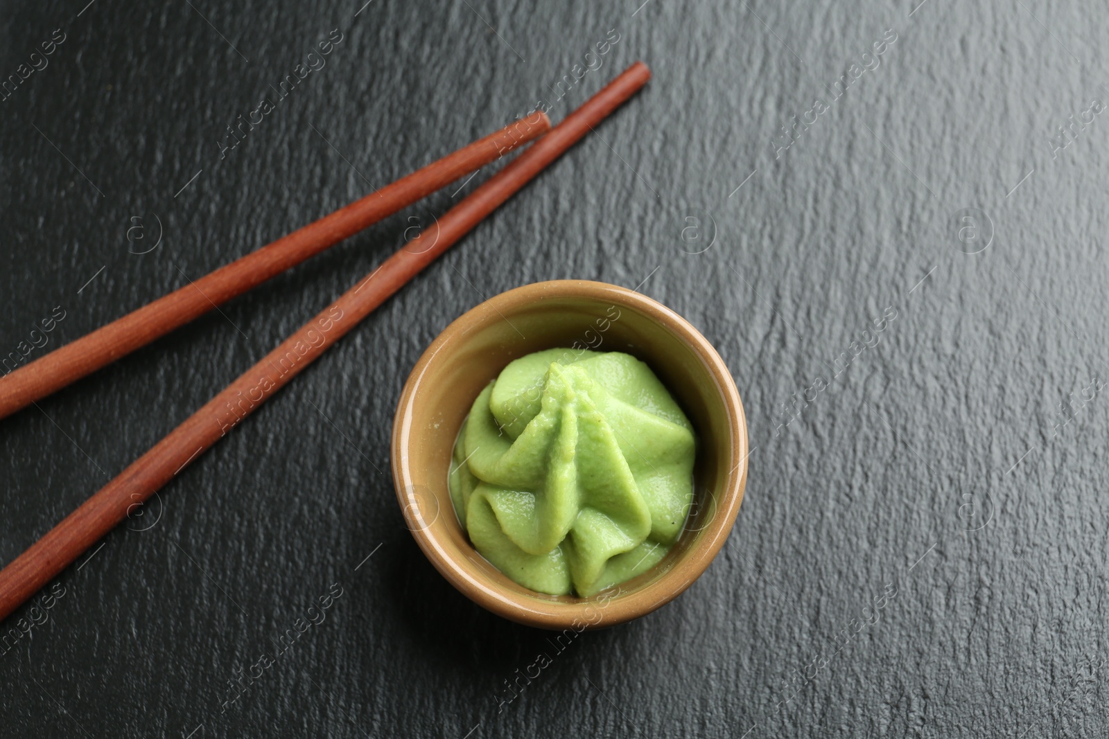 Photo of Hot wasabi paste in bowl and chopsticks on dark textured table, top view