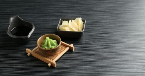 Photo of Hot wasabi paste, soy sauce and ginger on black textured table. Space for text