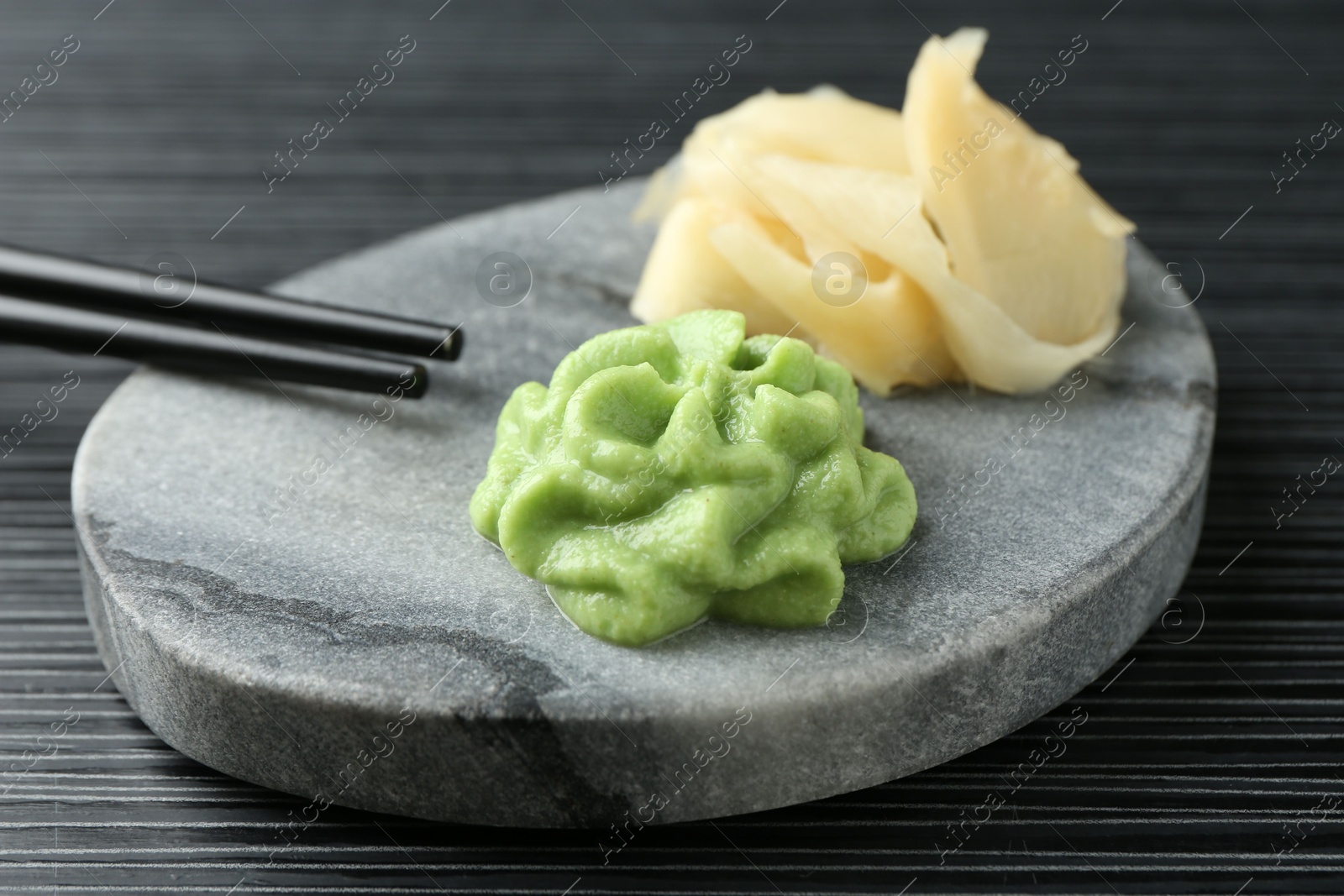 Photo of Marble board with hot wasabi paste, ginger and chopsticks on black textured table, closeup