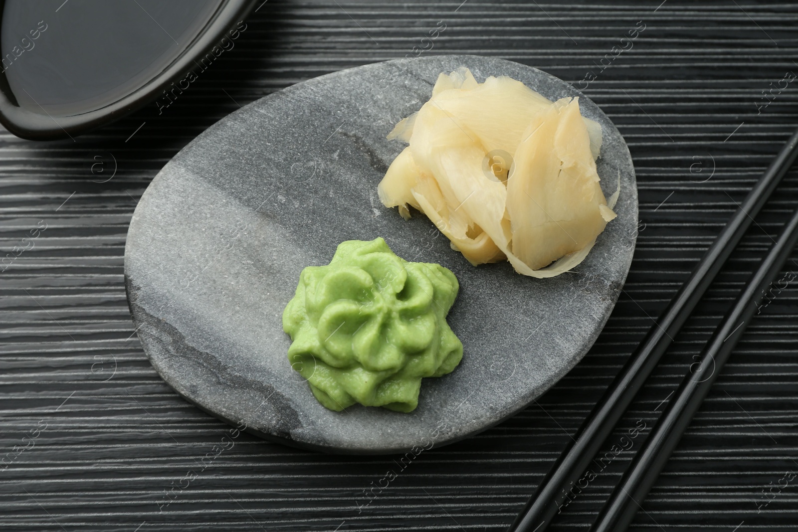 Photo of Hot wasabi paste, soy sauce, ginger and chopsticks on black textured table, flat lay