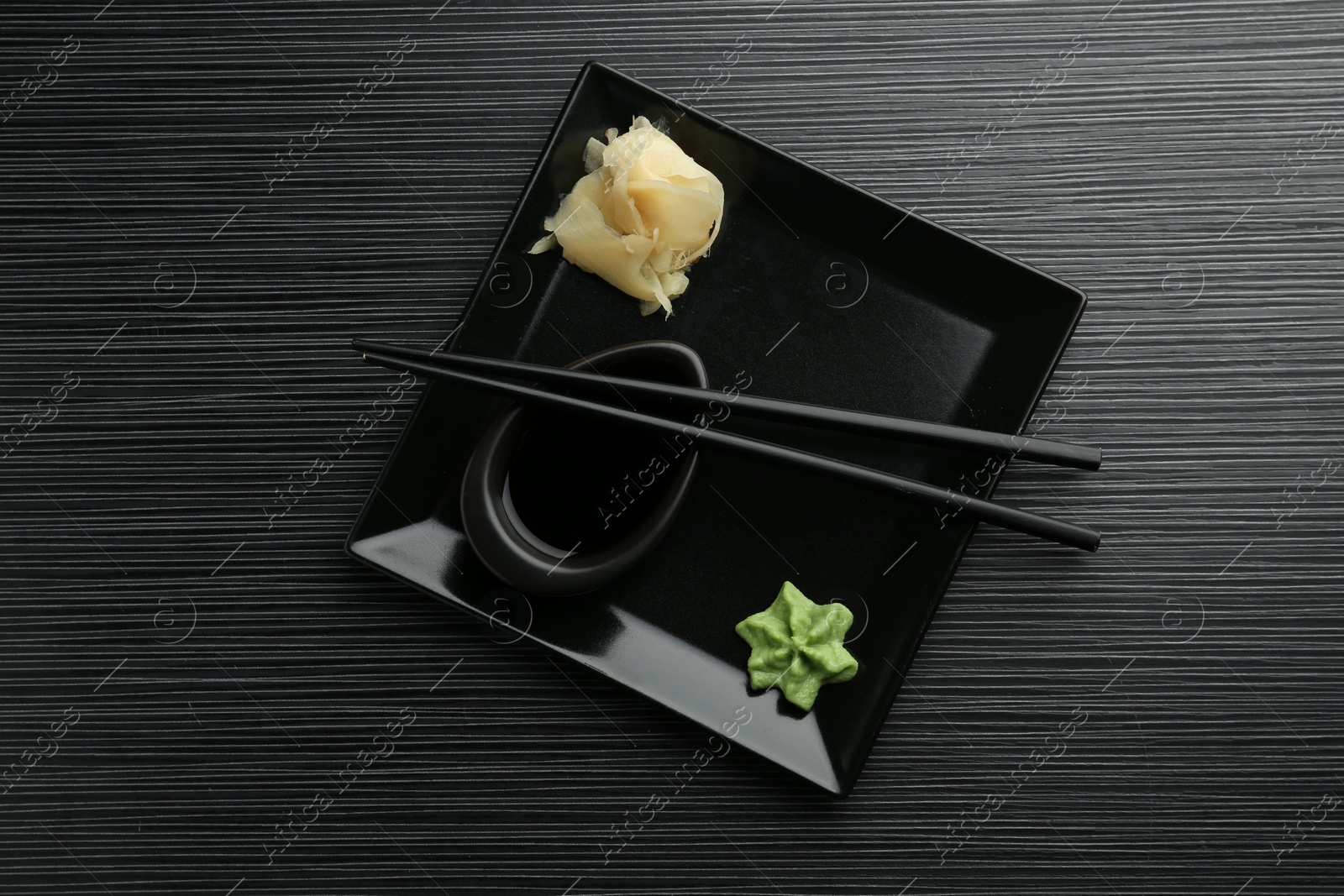 Photo of Hot wasabi paste, soy sauce, ginger and chopsticks on black textured table, top view