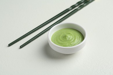 Photo of Hot wasabi paste in bowl and chopsticks on light textured table, closeup