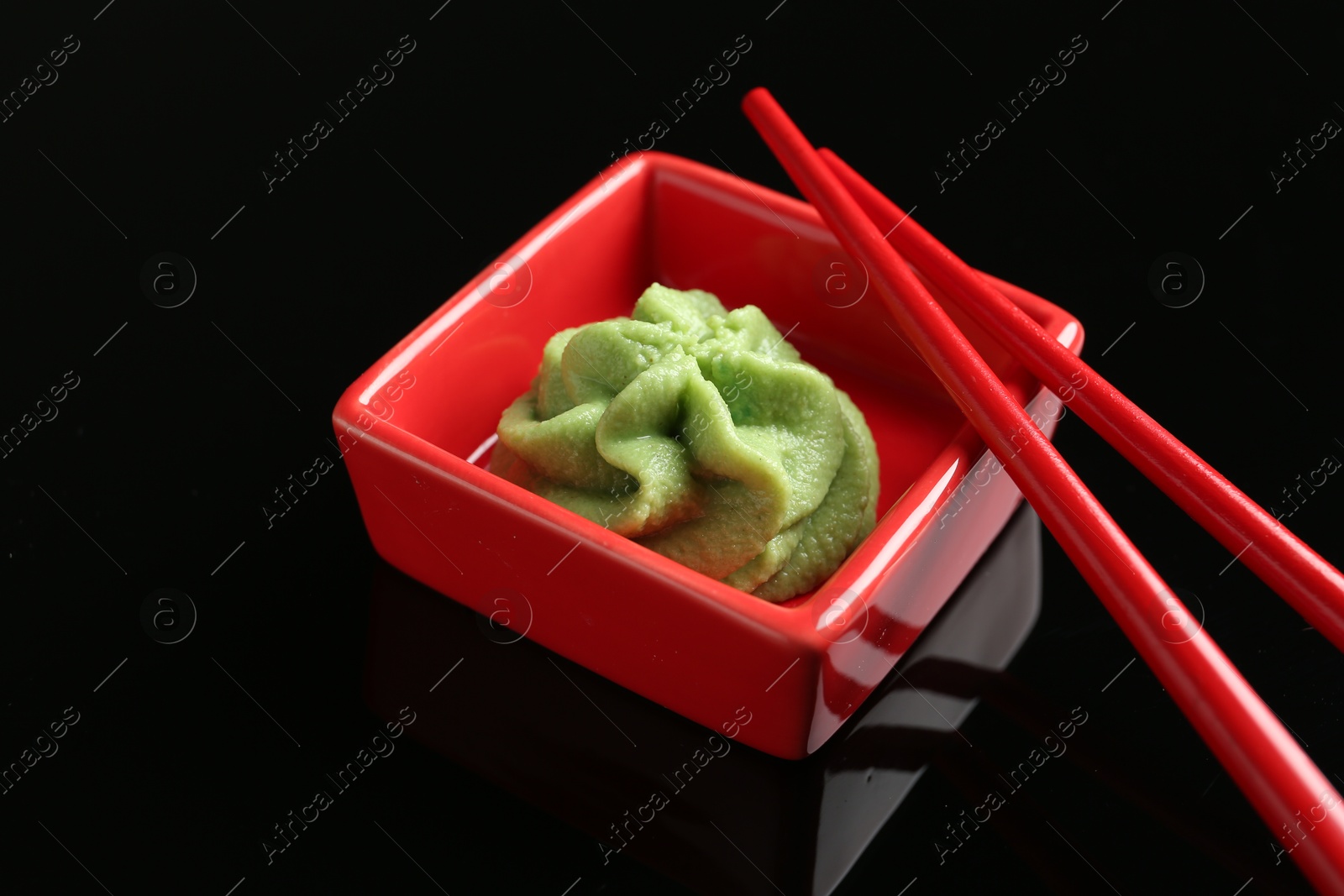 Photo of Hot wasabi paste in bowl and chopsticks on black mirror surface, closeup