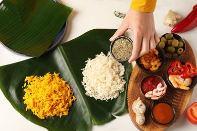 Photo of Woman holding bowl of cumin at white table with banana leaves and tasty food, top view