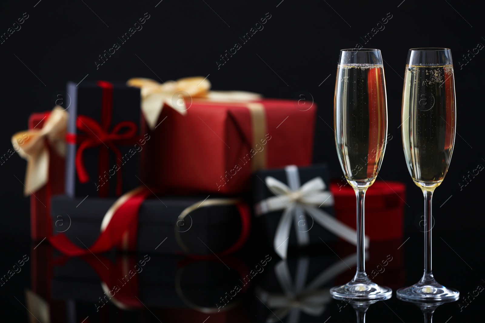 Photo of Delicious champagne in glasses and Christmas presents on black mirror surface, closeup. Space for text