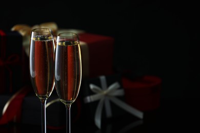 Photo of Delicious champagne in glasses and Christmas presents on black background, closeup. Space for text