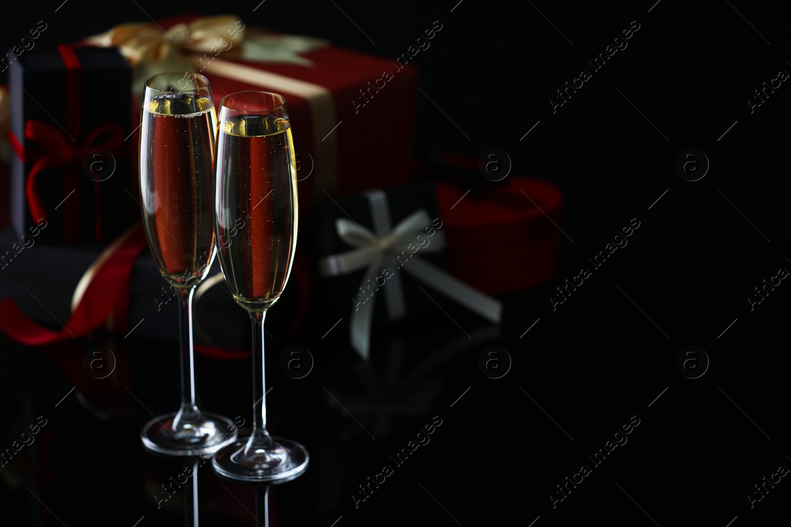 Photo of Delicious champagne in glasses and Christmas presents on black mirror surface, closeup. Space for text