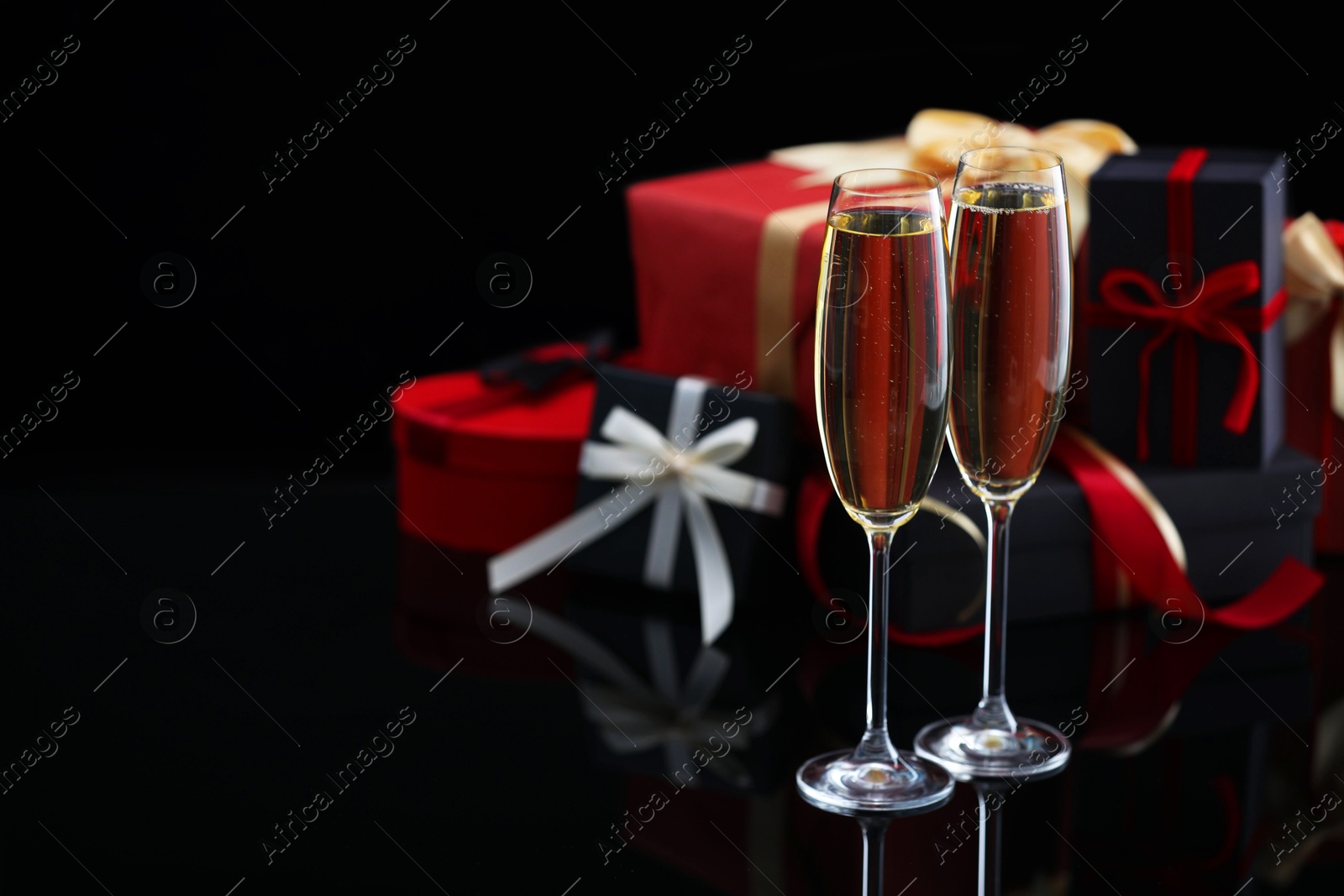 Photo of Delicious champagne in glasses and Christmas presents on black mirror surface, closeup. Space for text