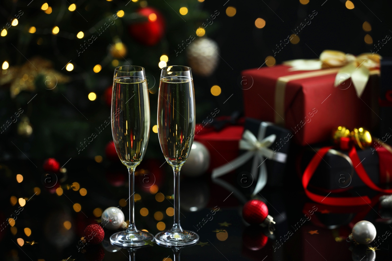 Photo of Delicious champagne in glasses and Christmas decor on black mirror surface against blurred lights