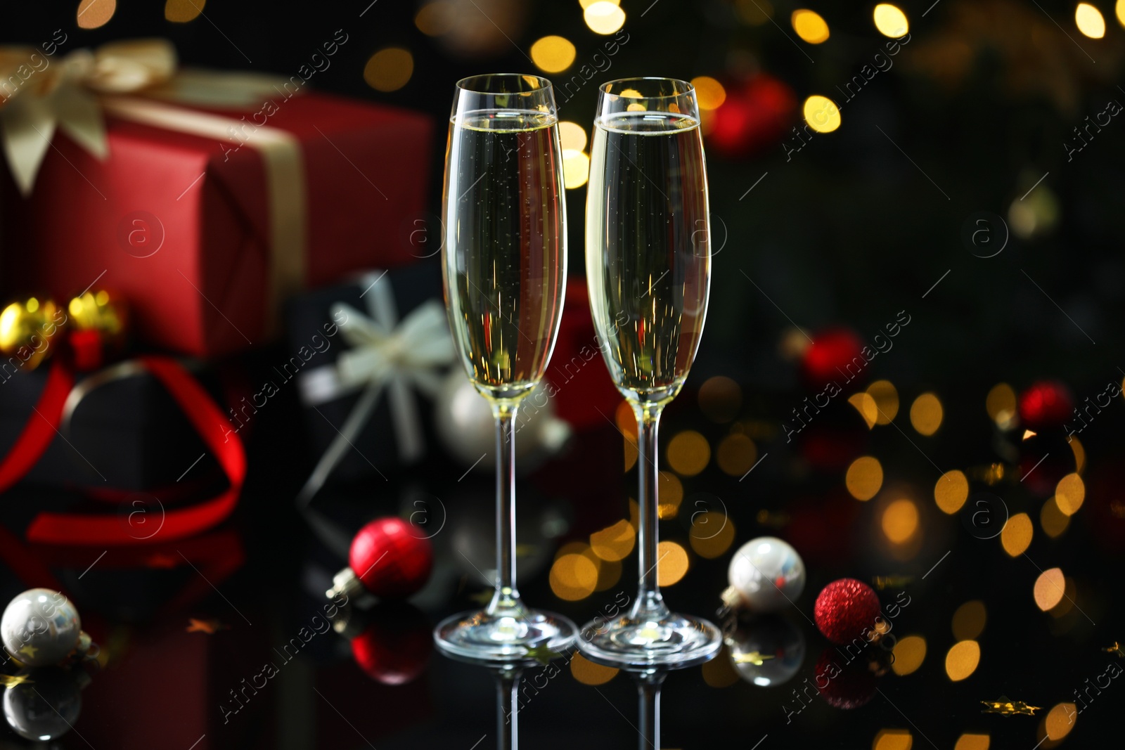 Photo of Delicious champagne in glasses and Christmas decor on black mirror surface against blurred lights, closeup