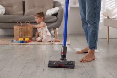 Photo of Young woman cleaning floor with cordless vacuum cleaner while her daughter playing with toys at home, closeup