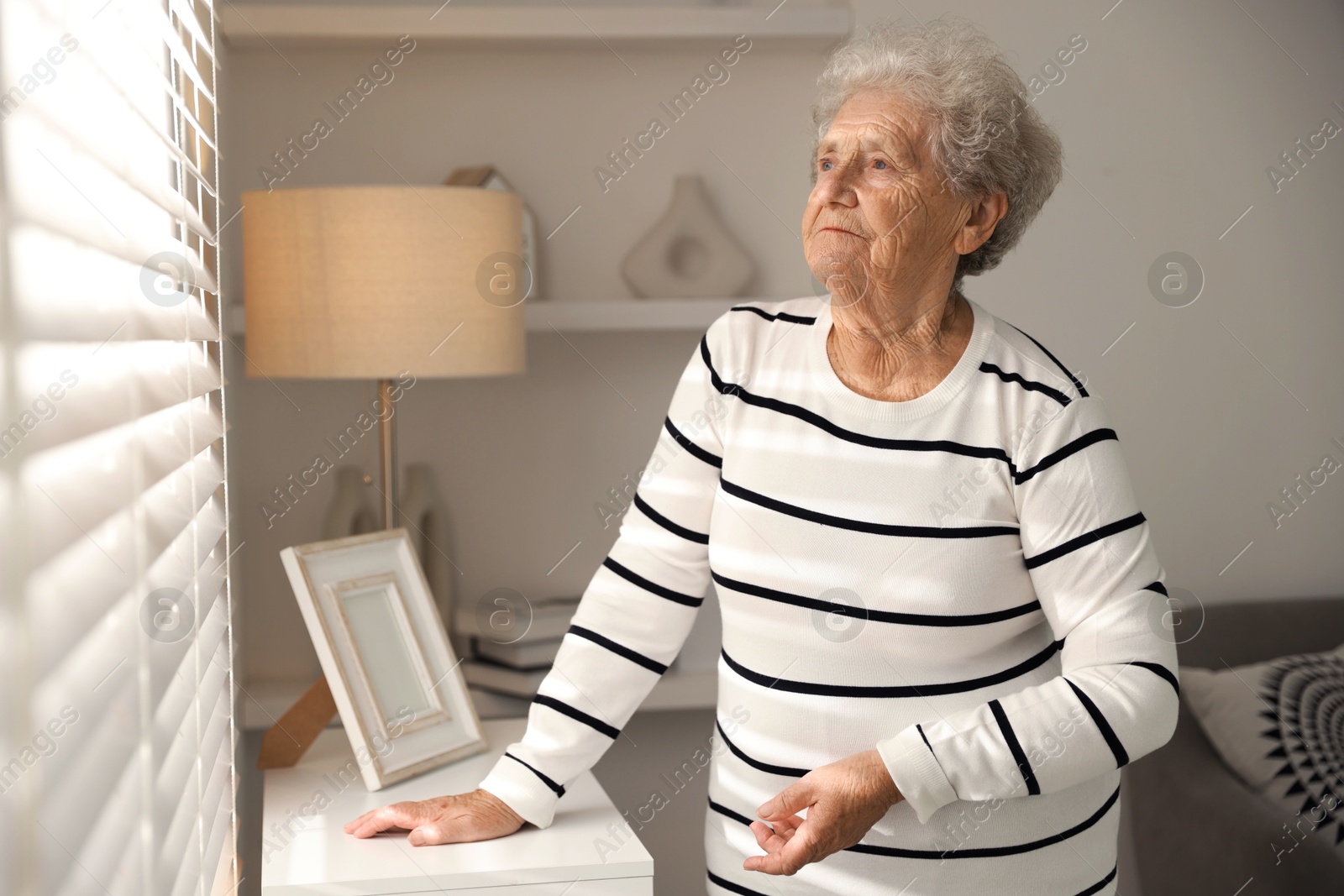 Photo of Loneliness concept. Sad senior woman looking through window blinds at home