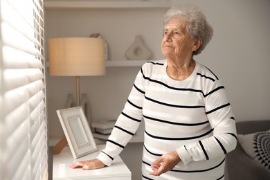 Photo of Loneliness concept. Sad senior woman looking through window blinds at home