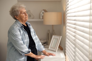 Photo of Loneliness concept. Sad senior woman looking through window blinds at home