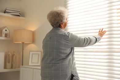 Photo of Loneliness concept. Sad senior woman looking through window blinds at home