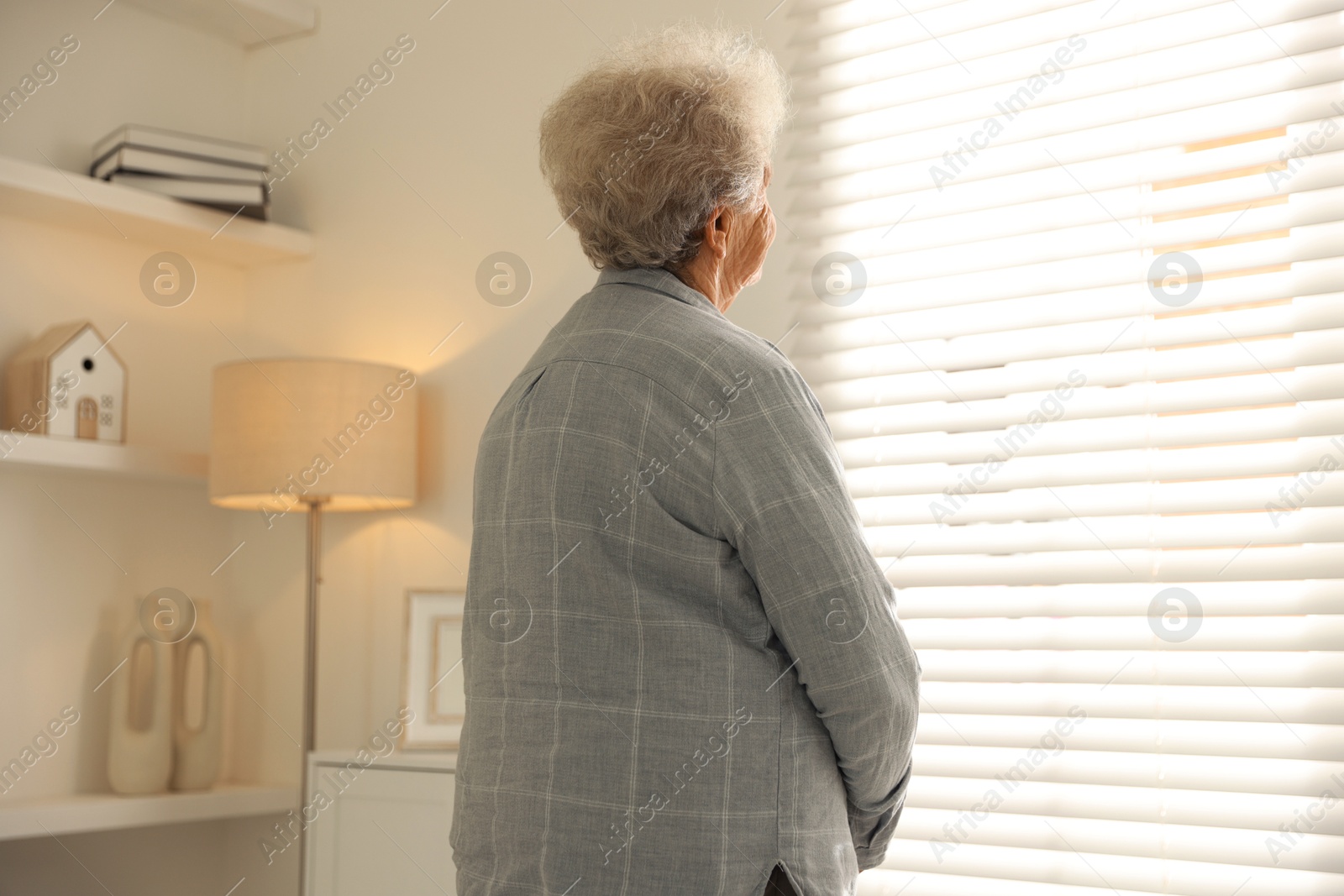 Photo of Loneliness concept. Sad senior woman looking through window blinds at home