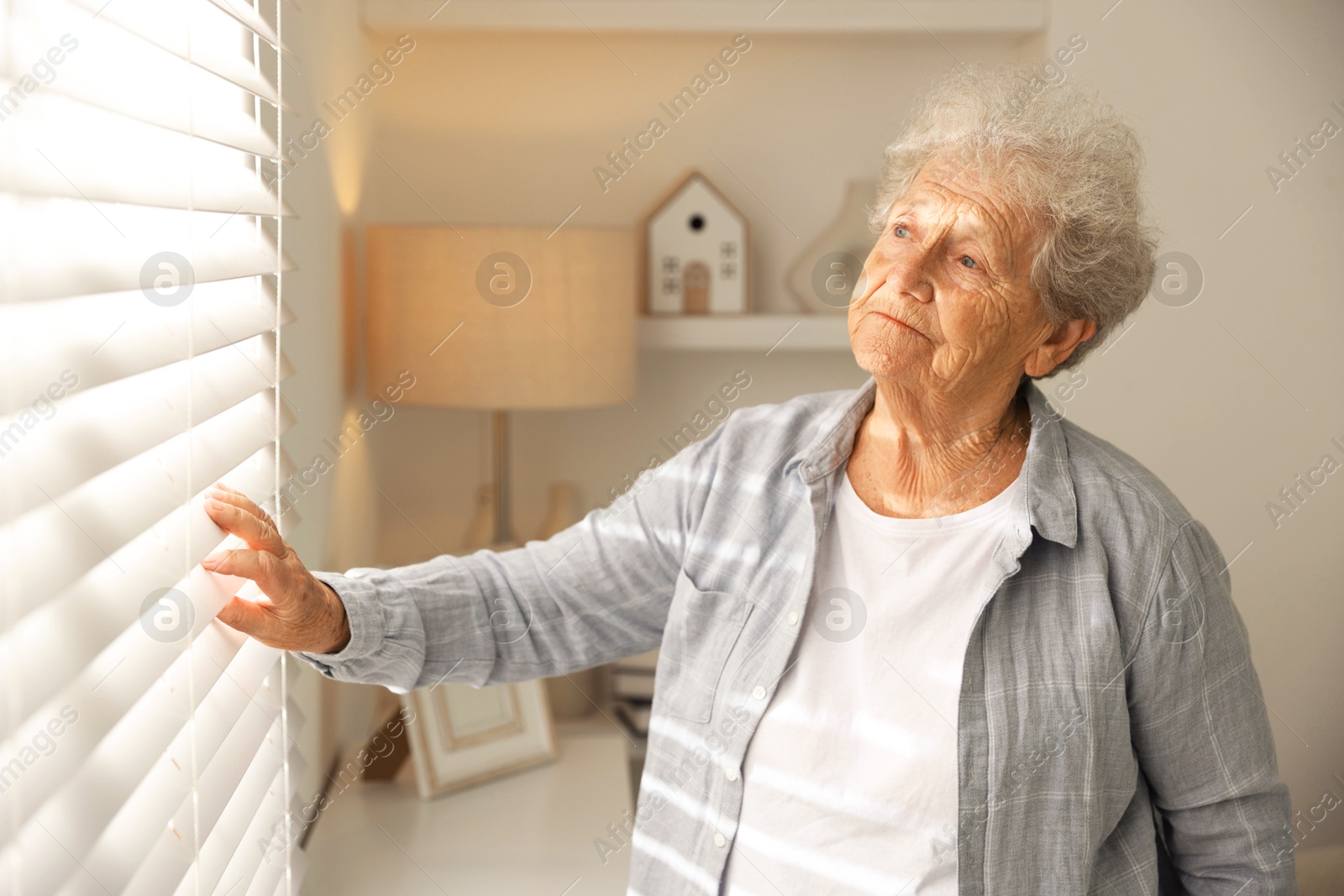 Photo of Loneliness concept. Sad senior woman looking through window blinds at home