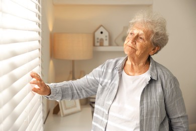 Photo of Loneliness concept. Sad senior woman looking through window blinds at home