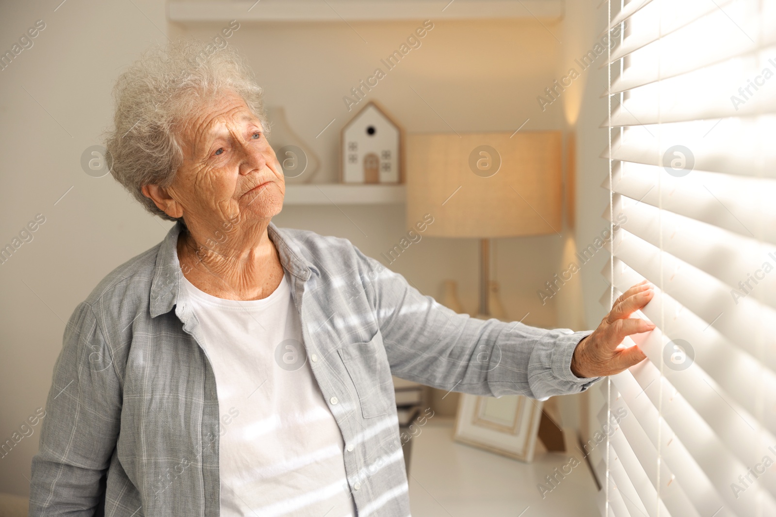 Photo of Loneliness concept. Sad senior woman looking through window blinds at home